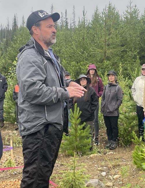 Carl Pollard in a forest field. He is in a grey jacket with black pants. He is surrounded by trees and students listening to him intently.